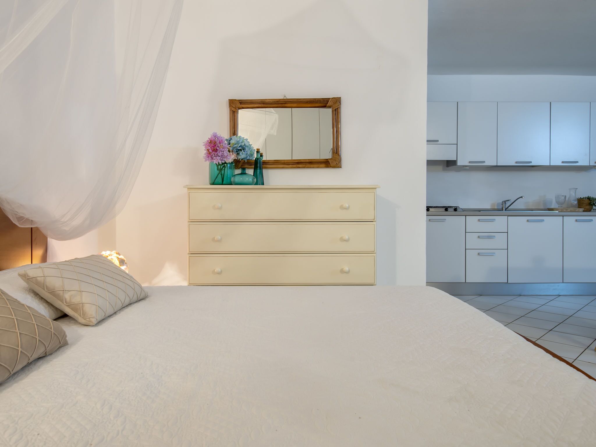 Un dormitorio con cama, una mesita de noche con un florero, un espejo y una kitchenette al fondo.