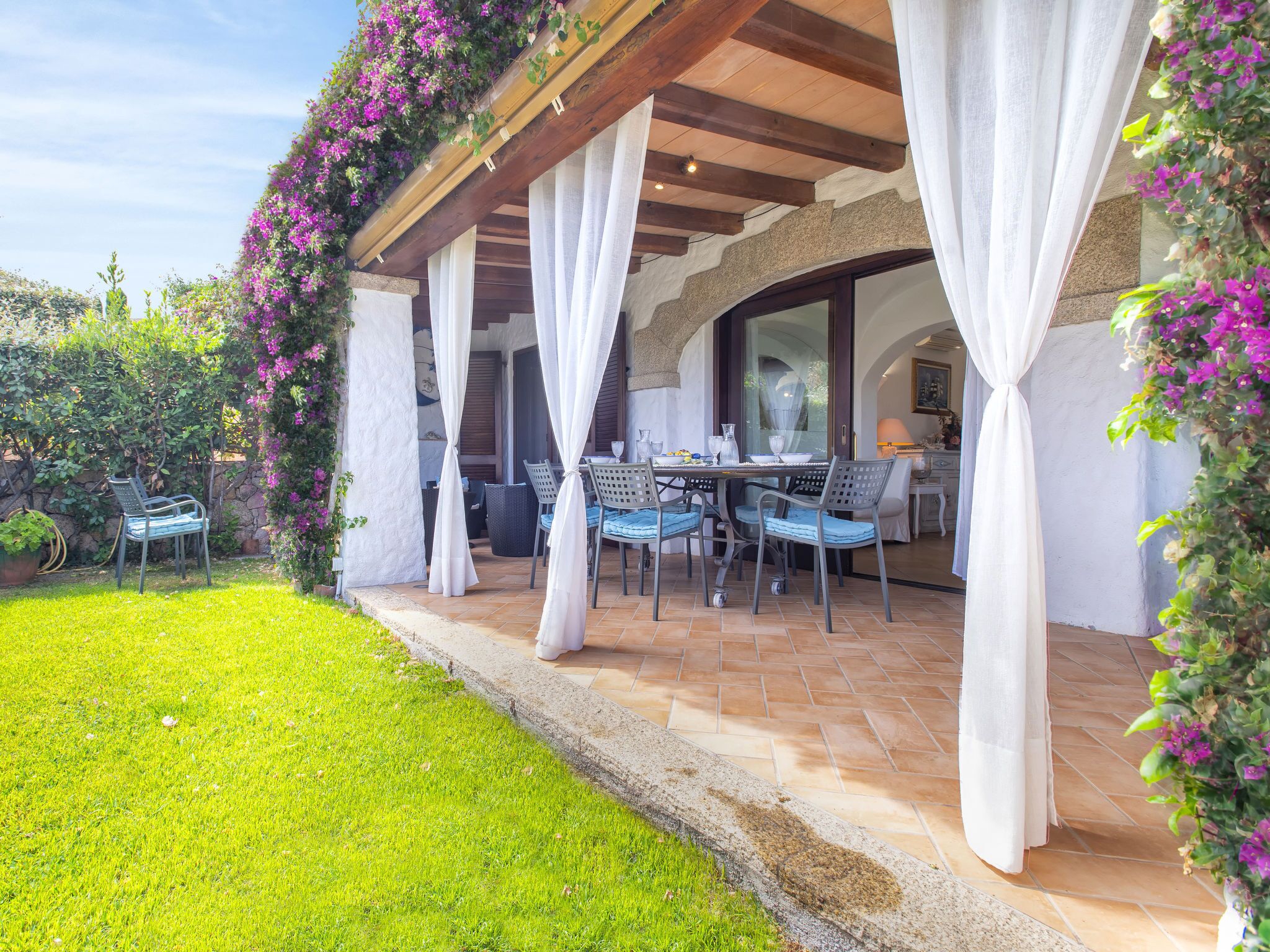 En udendørs terrasse med borde og stole, omgivet af en stenvæg og overdækket af et træpergola.