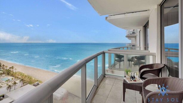 A balcony with a view of the ocean, furnished with wicker chairs and a table with bottles. - 1BR Apartment with beautiful view (Sunny Isles Beach)