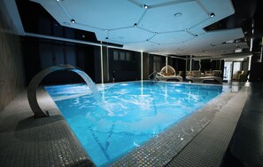 A modern indoor swimming pool with a clear water feature, surrounded by lounge chairs and a contemporary ceiling design.