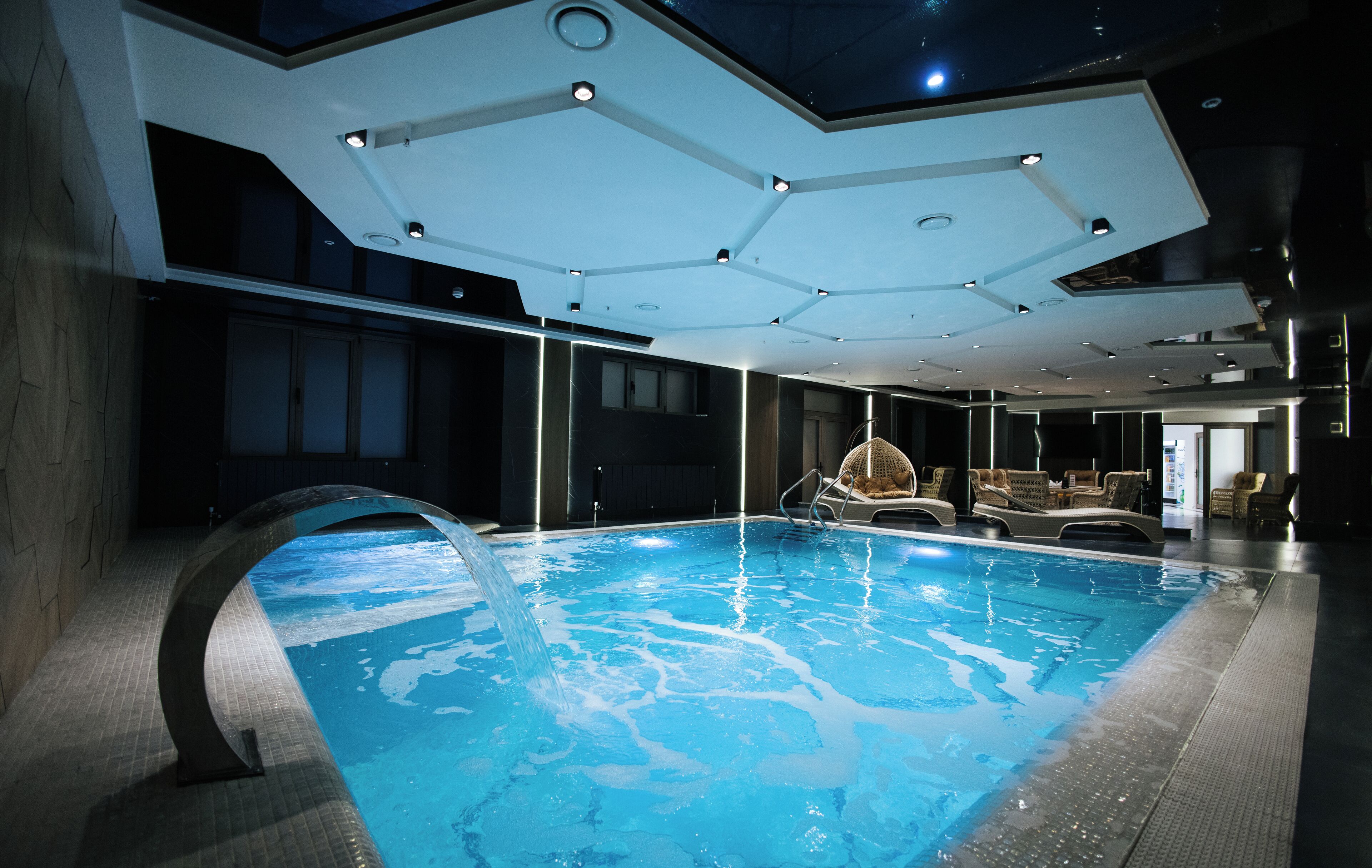 Una piscina interior con diseño moderno, que incluye una estructura de techo única y un área de descanso con sillas.