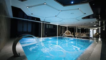 Una piscina interior con diseño moderno, que incluye una estructura de techo única y un área de descanso con sillas.