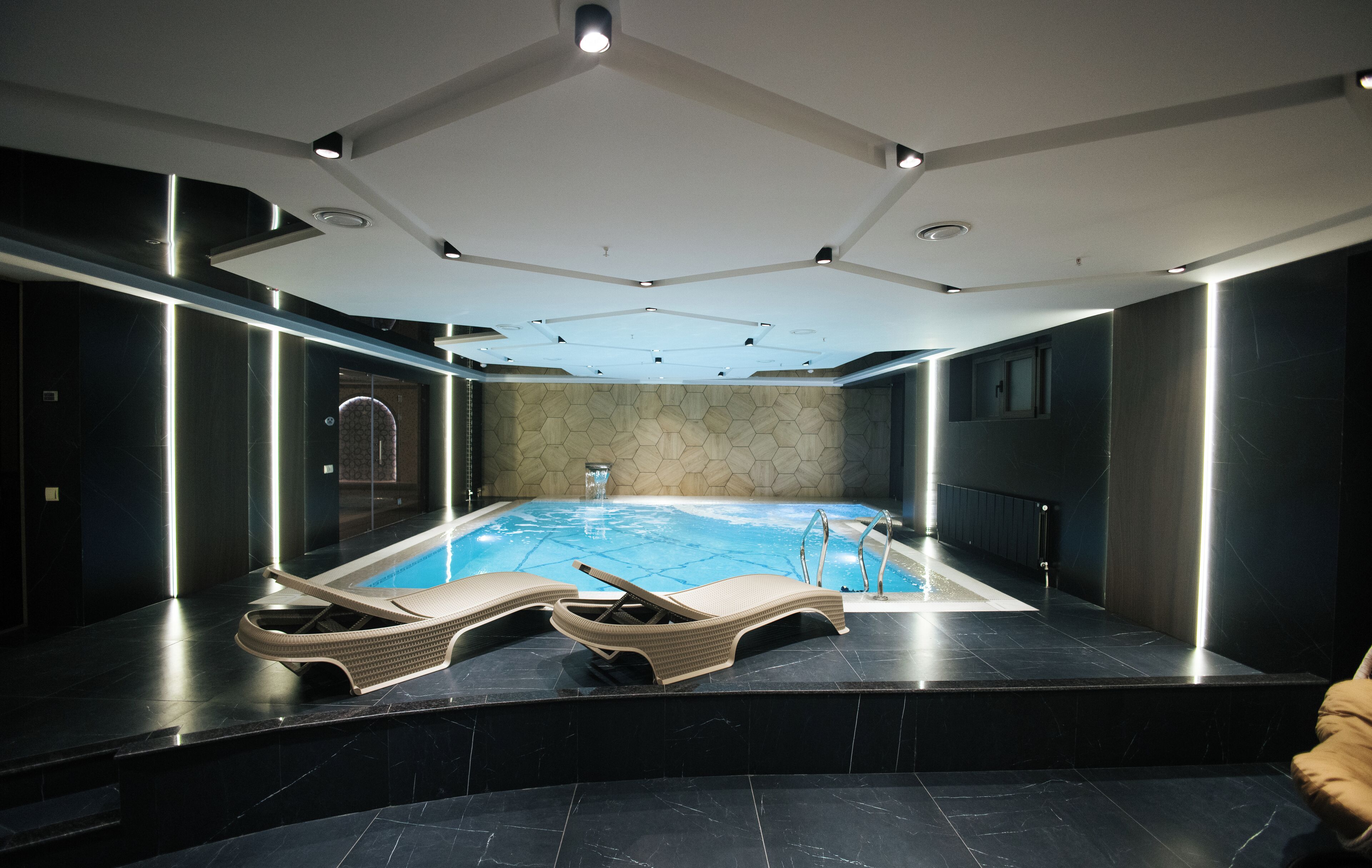 Une piscine intérieure moderne dotée de deux chaises longues, d’une entrée avec porte vitrée et d’un plafond à motifs hexagonaux.
