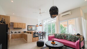 Uma cozinha moderna com armários de madeira, área de jantar com mesa e cadeiras, um sofá rosa e uma mesa de centro preta com frutas.