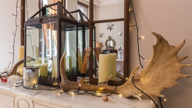 A fireplace mantel with a mirror, candles, and decorative items including a large antler and string lights.
