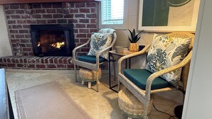 A cozy living room with a brick fireplace, wicker chairs with cushions, a woven rug, and a framed welcome sign.