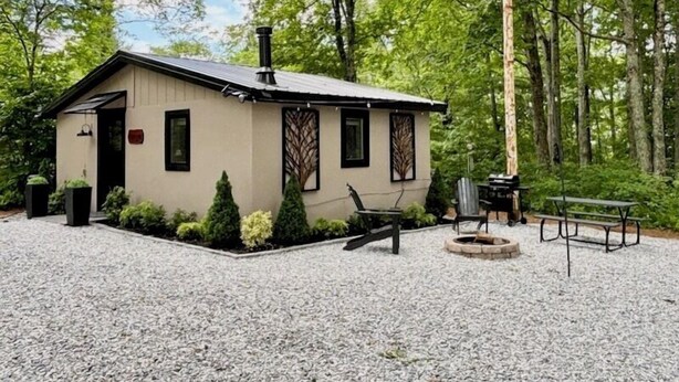A small cabin with a gravel driveway, a fire pit, and outdoor seating.