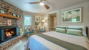 A bedroom with a fireplace, a bed with green and white bedding, a ceiling fan, and artwork on the walls.