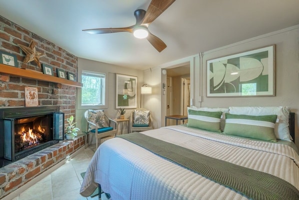 Une chambre avec une cheminée, un lit avec une literie verte et blanche, un ventilateur de plafond et des œuvres d’art accrochées aux murs.