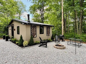 Une petite cabane avec une cheminée, un foyer extérieur, des sièges pour se détendre en plein air, le tout entouré d’arbres.