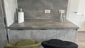 Een moderne keuken met een grijs aanrecht, twee barkrukken, een waterfles en een spuitbus.