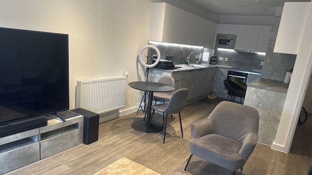 Een moderne woonkamer met een flatscreen televisie, een eethoek met een ronde tafel en stoelen, en een keuken met een magnetron en fornuis.