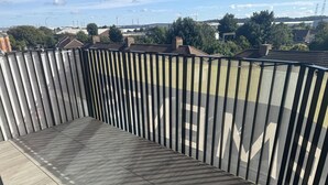 Een balkon met een metalen hekwerk en betegelde vloer, met uitzicht op een woongebied met huizen en bomen.