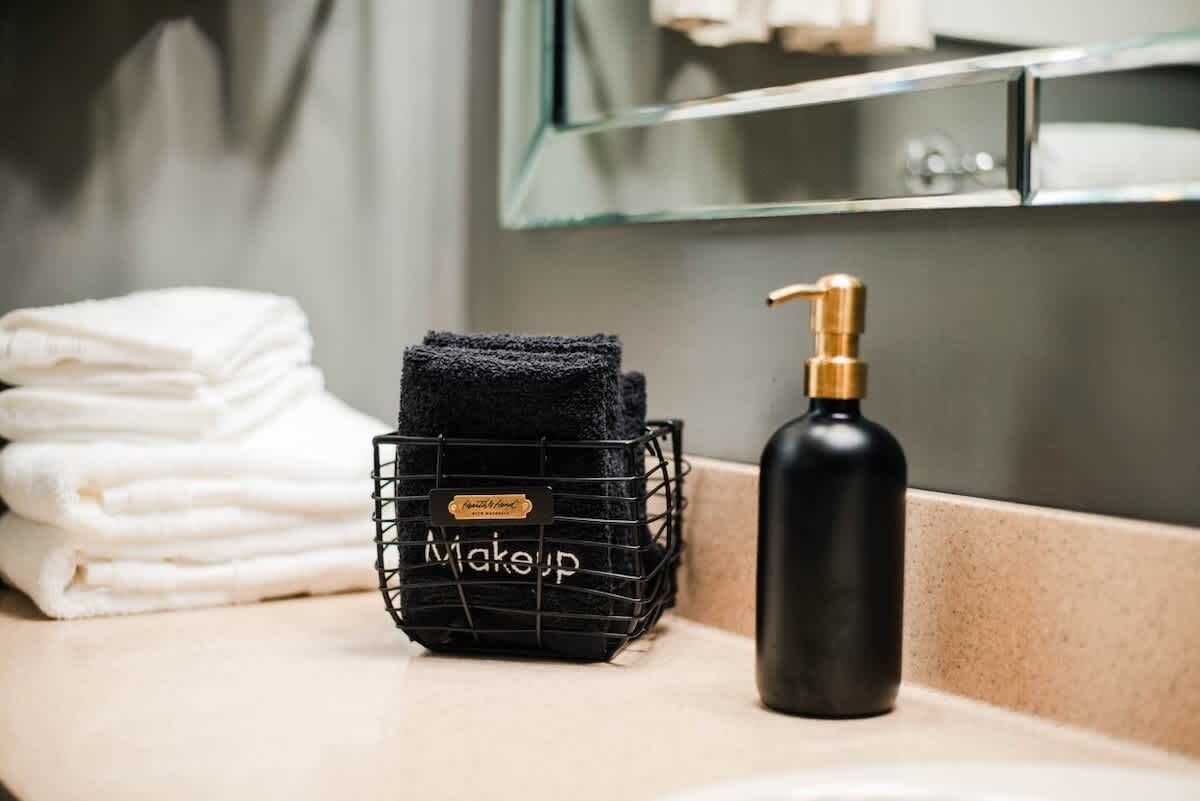 Ein Badezimmer-Waschtisch mit einem schwarzen Kosmetikkorb mit dem Aufdruck „Kosmetik“ und einer schwarzen Flasche neben ordentlich gefalteten weißen Handtüchern.
