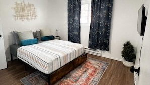 A bedroom with a bed, a ceiling fan, a window with curtains, a small plant, and a wall-mounted TV.