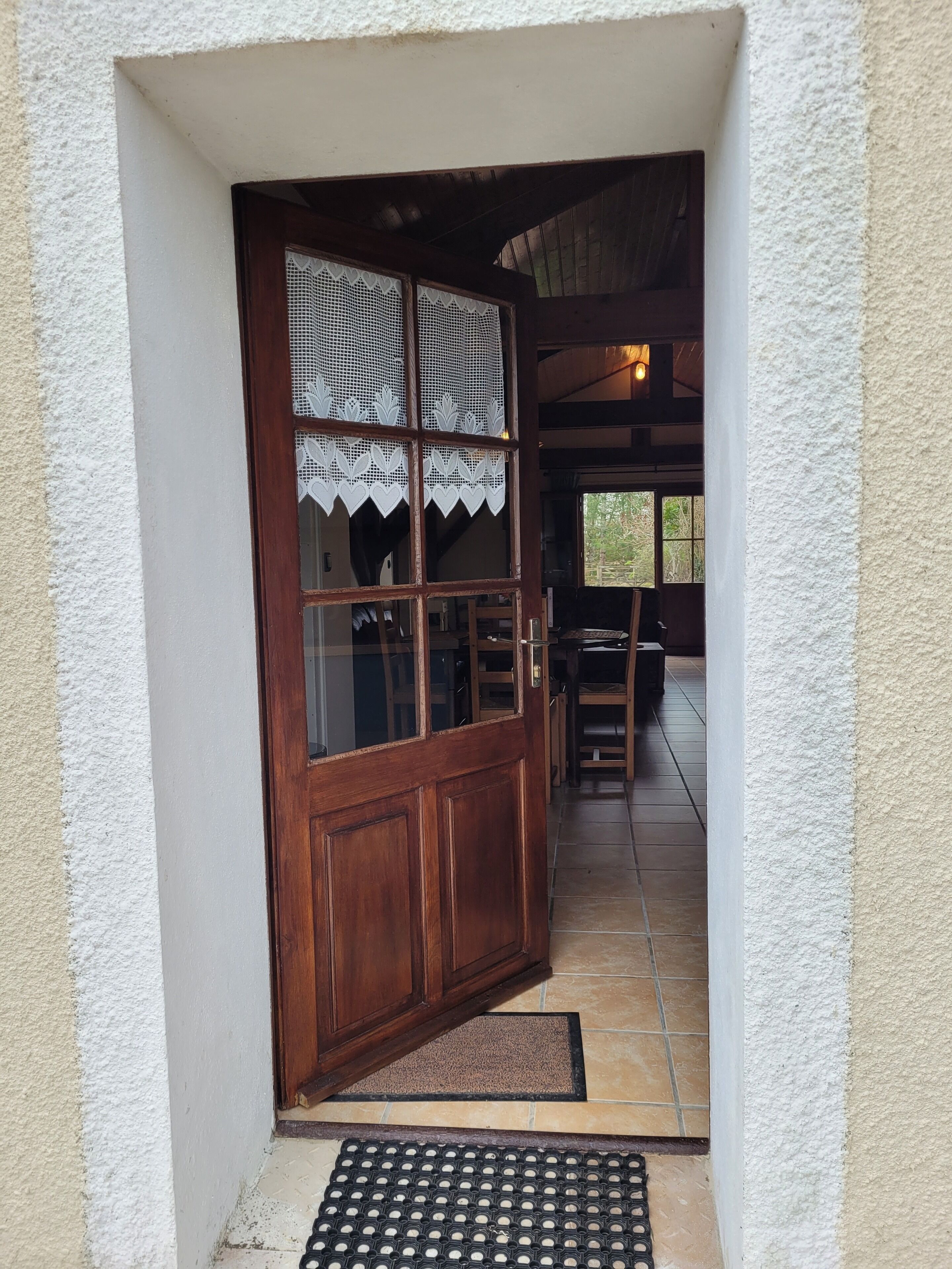 A wooden door with lace panels, a doormat, and a tiled floor leading to an interior space with a wooden bench and a table.