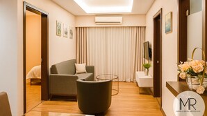 Una habitación de hotel moderna con sofá, una mesita redonda, una mesita auxiliar con un florero y una cama con edredón blanco, además de un amplio ventanal con cortinas.