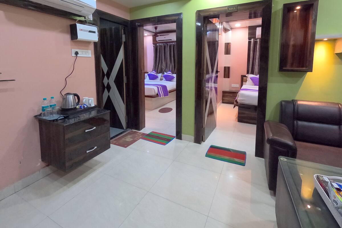 A hotel room with a bed, a desk, a chair, a sofa, an air conditioning unit, and a door leading to another room.