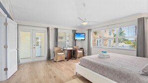 A hotel room with a bed, a desk with chairs, a television, and a view of the outside. - AMI Du Soleil | 1 Block 2 Beach, Big Heated & Shaded Saltwater Pool, Lanai, Grill, Walk to Restaurants, Cafes, Shops, Bars (Anna Maria)
