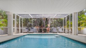 A swimming pool with clear blue water, surrounded by lounge chairs and a covered area with large windows overlooking lush greenery. - AMI Du Soleil | 1 Block 2 Beach, Big Heated & Shaded Saltwater Pool, Lanai, Grill, Walk to Restaurants, Cafes, Shops, Bars (Anna Maria)