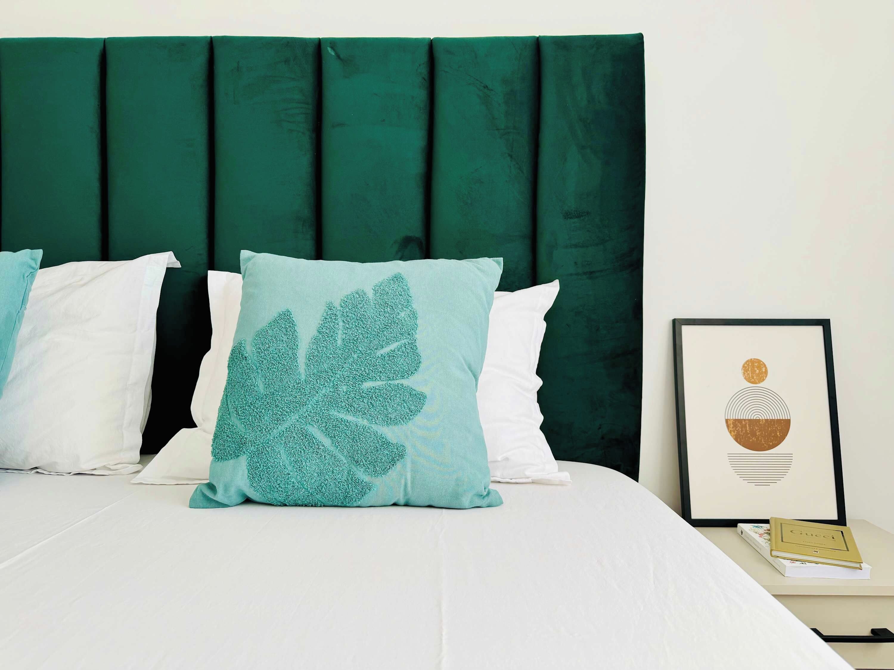 A bed with a dark green headboard, white bedding, and a single teal pillow with a leaf pattern. A framed picture is visible on the nightstand beside the bed.