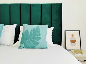 Ein Bett mit dunkelgrüner Kopfteil, weißer Bettwäsche und einem einzelnen, blaustürkisenen Kopfkissen mit Blätermotiv. Auf dem Nachttisch neben dem Bett ist ein gerahmtes Bild zu sehen.