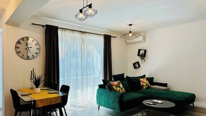 Een moderne woonkamer met een groene bank, een eettafel met stoelen, een ronde salontafel en een grote wandklok.