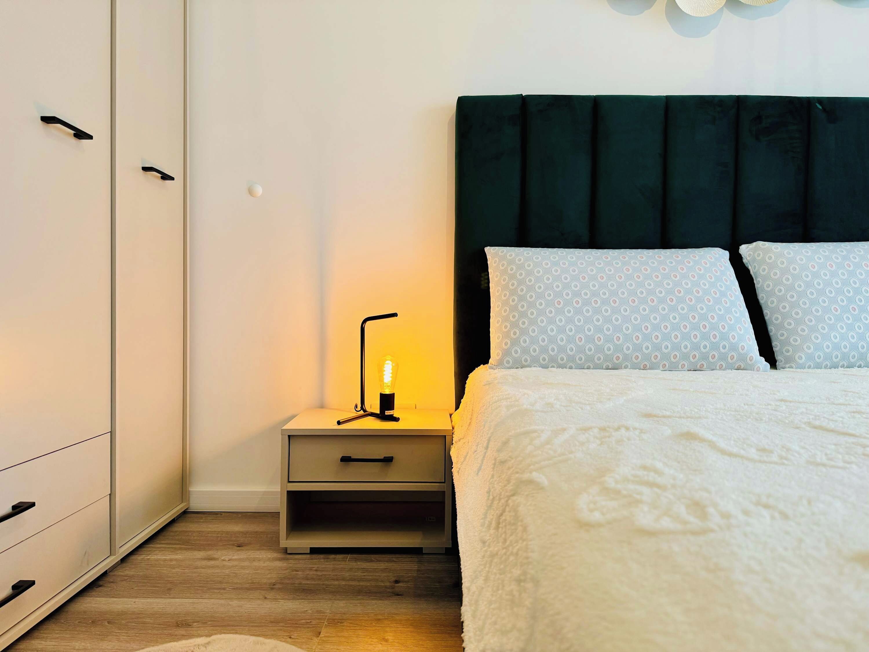 A bedroom with a bed, a nightstand, a lamp, a wardrobe, and a patterned pillow.