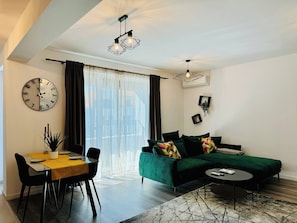 Una sala de estar moderna con un sofá verde, una mesa de comedor con sillas, una mesa redonda de centro y un gran reloj de pared.