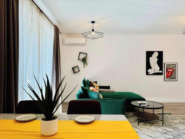 Una sala moderna con un sofá verde, una mesa de comedor con mantel amarillo y una mesita redonda. Hay dos cuadros en la pared y una planta en maceta sobre la mesa.
