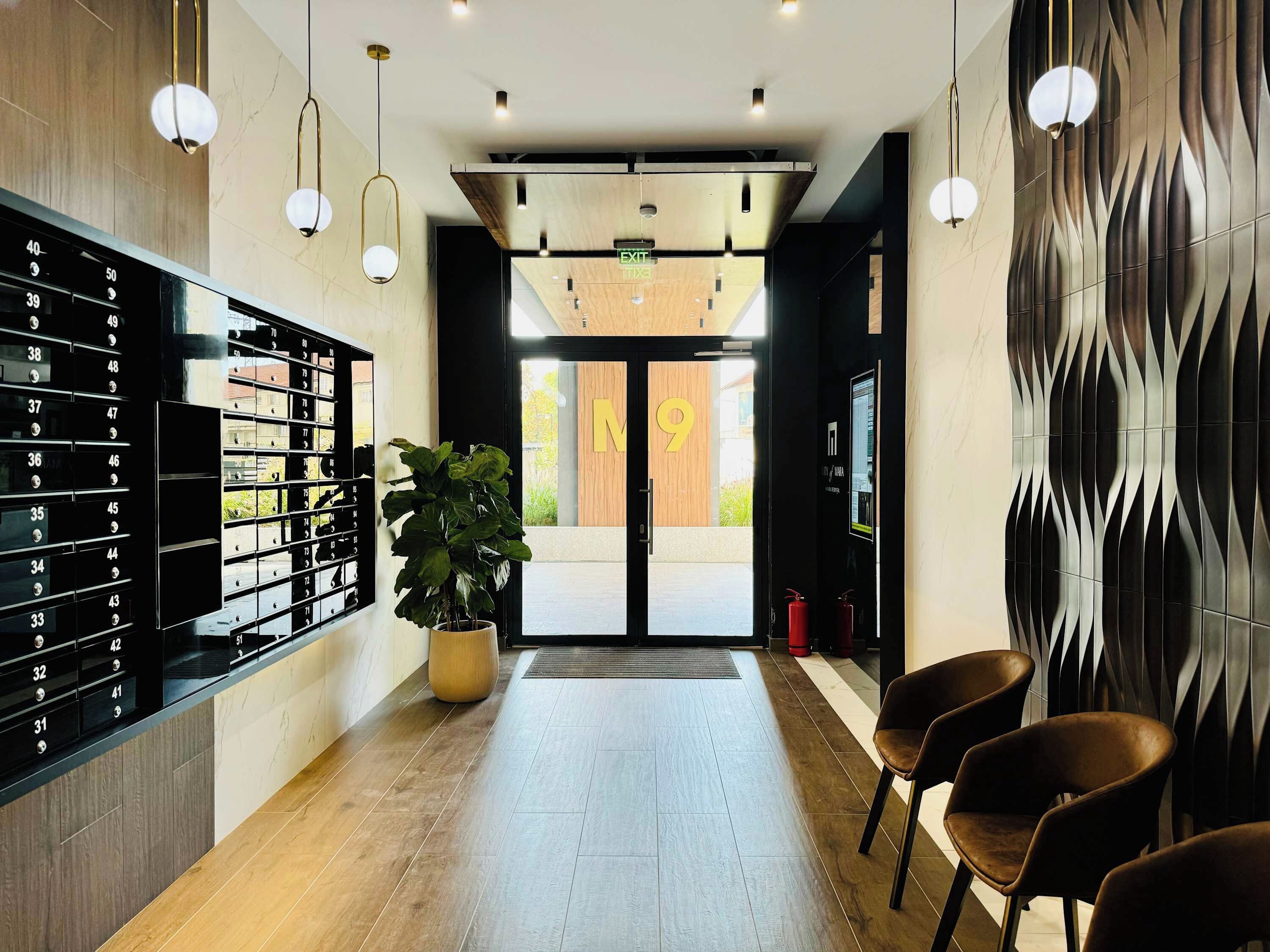 Een hotel lobby met een receptiebalie, een grote potplant, een brandblusser en een muur met een geometrisch patroon.