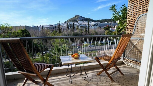 A balcony with two wooden chairs, a small table with fruits and a glass, and a view of a cityscape with a hill. - Lecabetus view apartment (Athina)