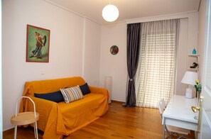 A living room with a sofa, a dining table, and a chair. There is a painting on the wall and a curtain covering the window.
