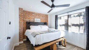 Ein Schlafzimmer mit einer Wand in Ziegeloptik, einem Bett mit weißer Bettwäsche, einem Deckenventilator und einem großen Fenster mit Vorhängen.