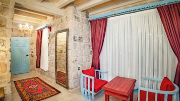 Une pièce aux murs de pierre, avec une porte bleue, un miroir, un rideau rayé de rouge et de blanc, une chaise rouge et bleue, et une petite table en bois.