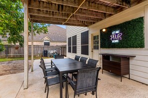 Une terrasse extérieure dotée d’une pergola en bois, d’une table à manger avec des chaises, d’un panneau lumineux au néon et d’un téléviseur fixé au mur.