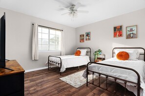 Une chambre avec deux lits simples, un ventilateur de plafond, une fenêtre avec des rideaux et deux tableaux accrochés au mur.