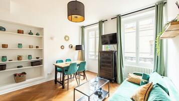 Een woonkamer met een bank, een glazen salontafel, een eettafel met stoelen, een televisie en een boekenkast.