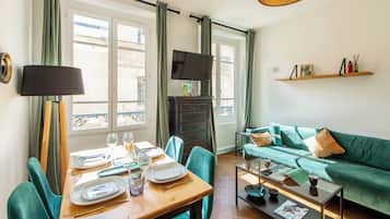 Een eetgedeelte met een tafel voor vier personen, een bank, een televisie en een plank met boeken en decoratieve objecten.
