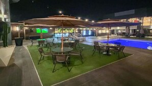 Un espace extérieur avec piscine, des tables et des parasols, entouré d’un petit muret et d’un bâtiment doté d’une enseigne lumineuse.