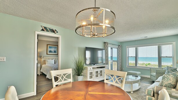 A spacious living room with a wooden dining table, white chairs, a chandelier, a television, and a view of the beach through large windows. - Dog Friendly-Sandy Dunes-Sleeps 8-3br/2b-Oceanfront-WIFI (North Myrtle Beach)