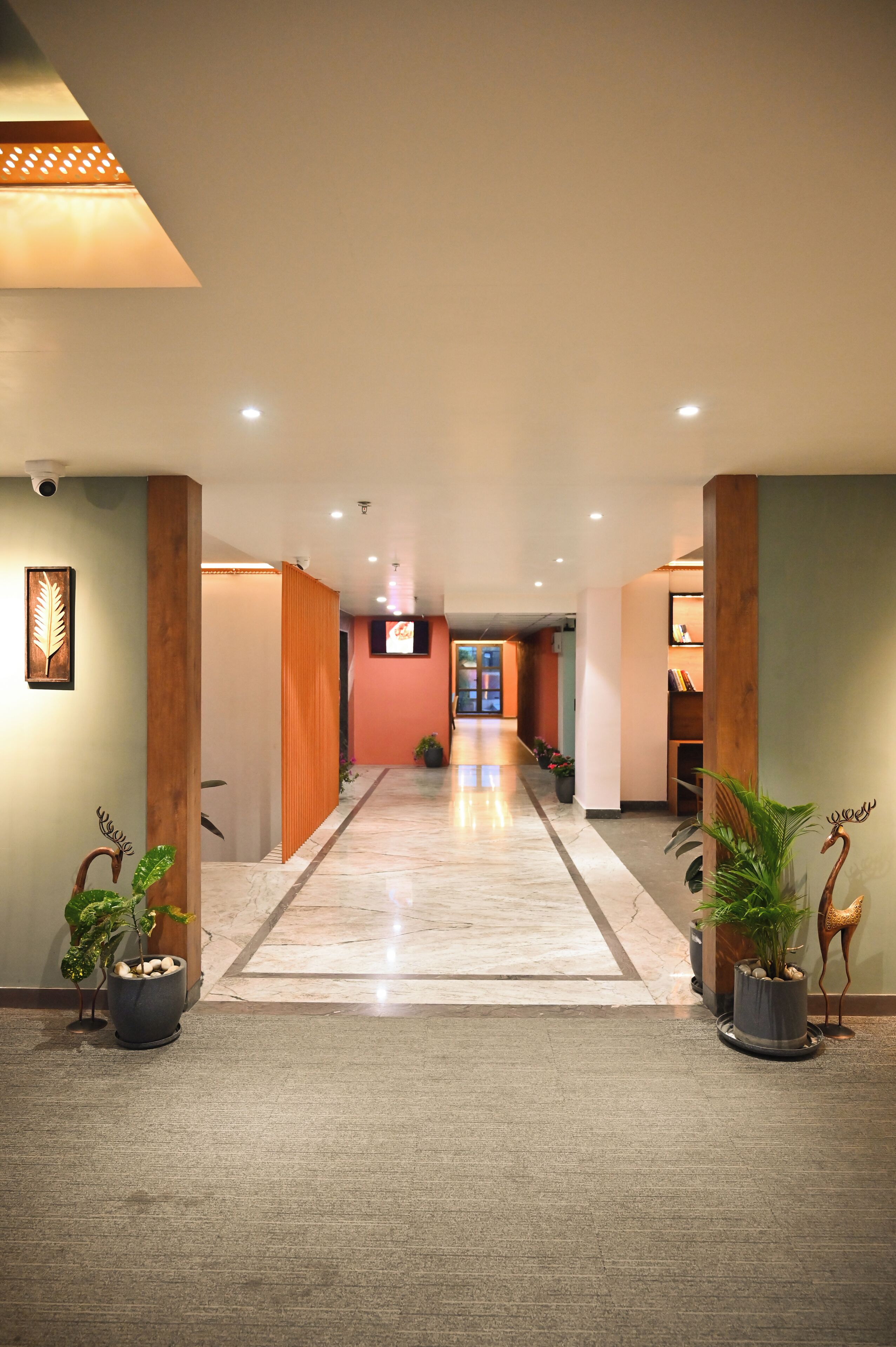 A hotel corridor with a marble floor, wooden columns, potted plants, and a television screen.