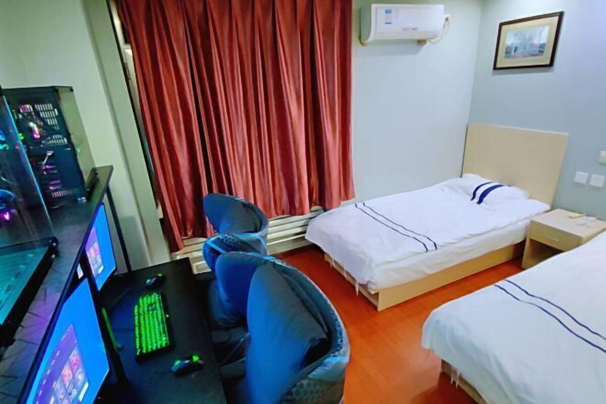 Hotelkamer met een bureau, twee bedden, een stoel, een rood gordijn en een airconditioner aan de muur.