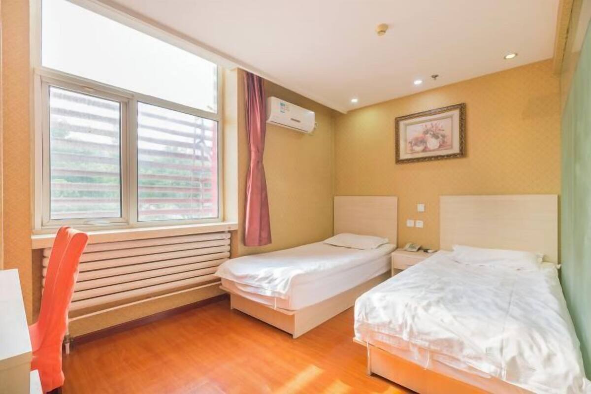 Een kamer met twee bedden, een raam met zonwering, een airconditioningapparaat, een lijst met een afbeelding aan de muur en een stoel.