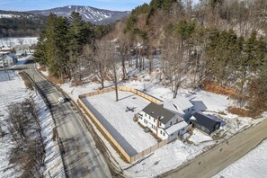 Área residencial com uma casa rodeada por neve, uma estrada e uma encosta arborizada.