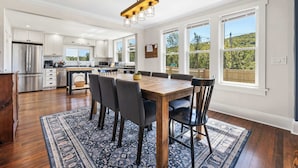 A modern kitchen with white cabinets, a wooden dining table, and a large window overlooking a green landscape.