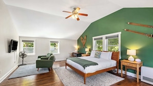 A bedroom with a sloped ceiling, a bed, a desk, a chair, a fan, and a window.