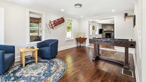 A spacious room with hardwood flooring, a foosball table, blue armchairs, a coffee table with board games, and a large window with a view of greenery.