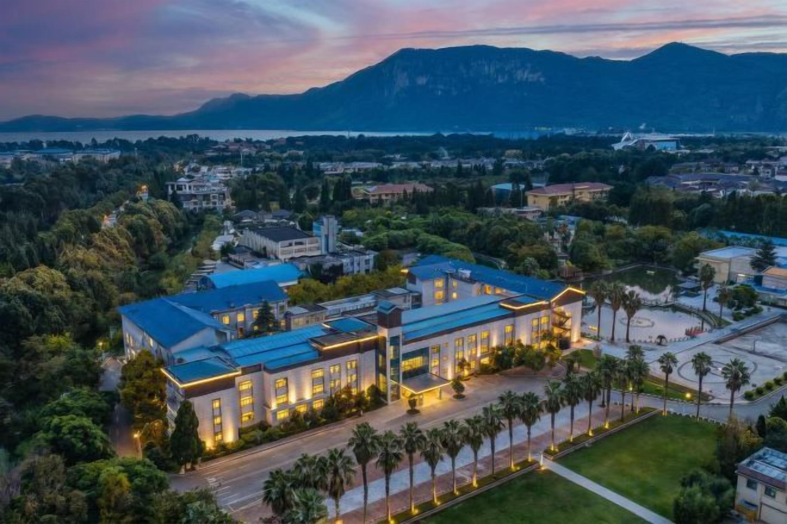 A large building with a blue roof, surrounded by greenery and palm trees, with a mountainous backdrop.