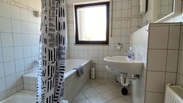 Un bany amb banyera, cortina de dutxassa, finestra, rentapans i radiador.